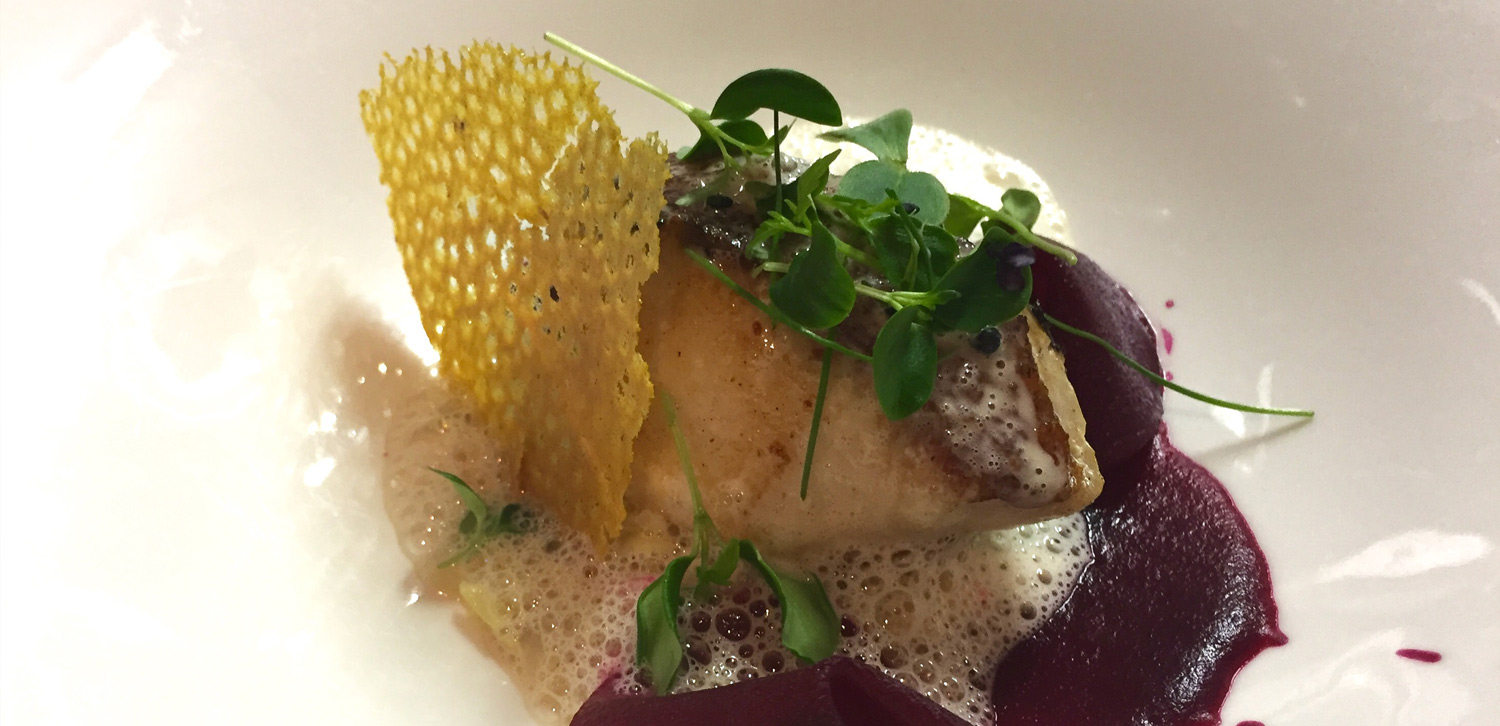 A gourmet dish featuring a delicately seared piece of fish, garnished with microgreens, foam, and a crisp golden lattice, accompanied by a deep purple beet puree.