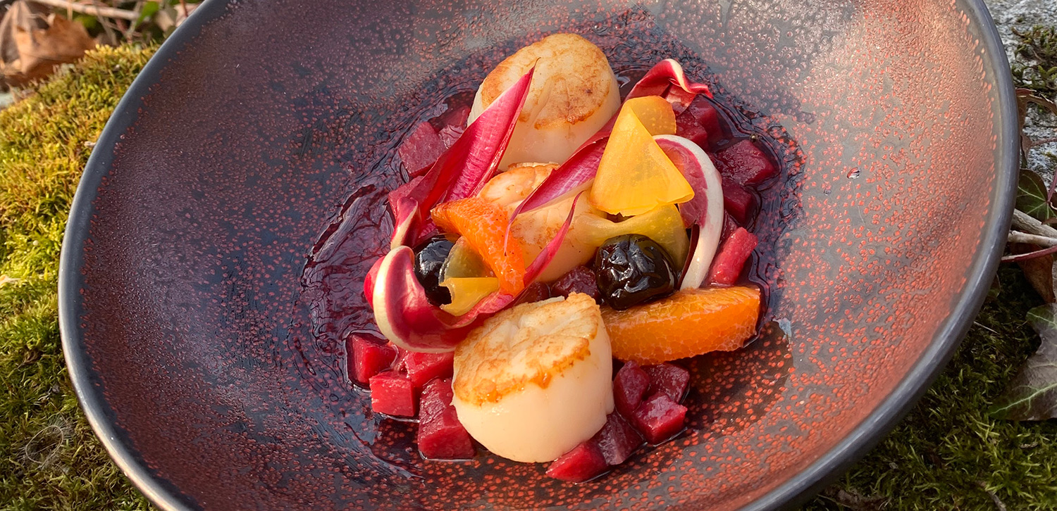 A beautifully plated gourmet dish featuring seared scallops with vibrant garnishes, including red and yellow root vegetables, olives, and endive leaves, served on a textured dark plate.