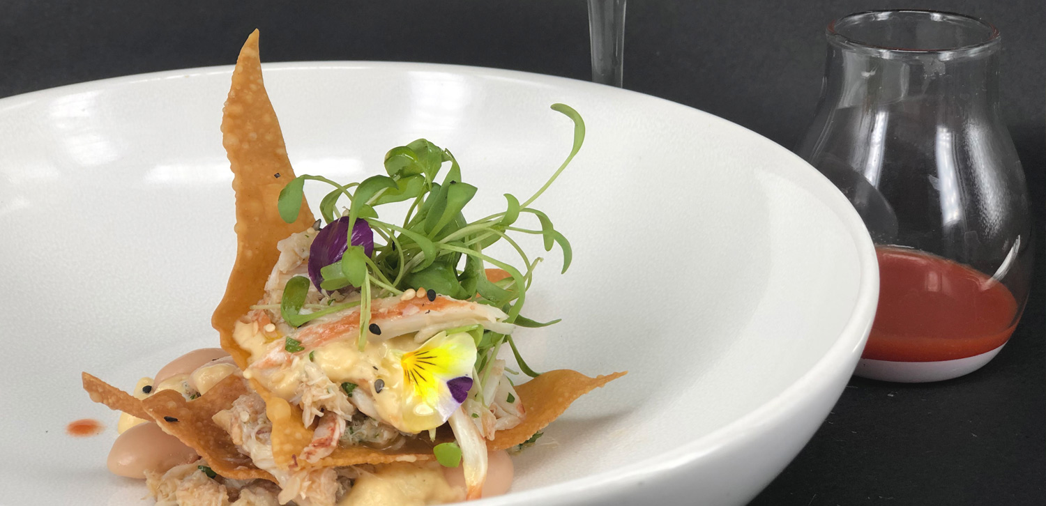 A refined gourmet dish with crab meat, crispy wonton pieces, microgreens, and edible flowers, served in a white bowl with a small glass of red sauce on the side.