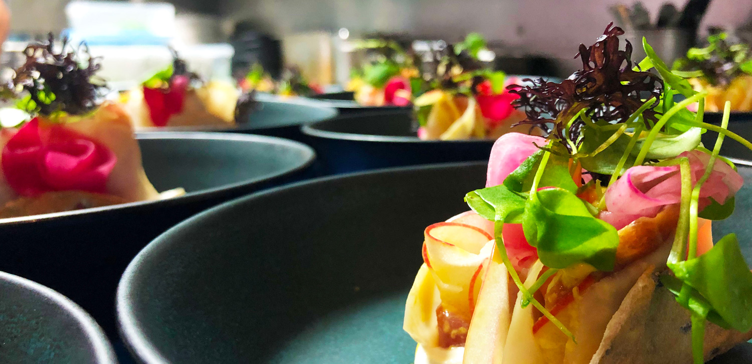 A close-up of a colorful gourmet appetizer with fresh greens, thinly sliced radishes, and a bright pink garnish, artfully arranged on multiple plates in a row.